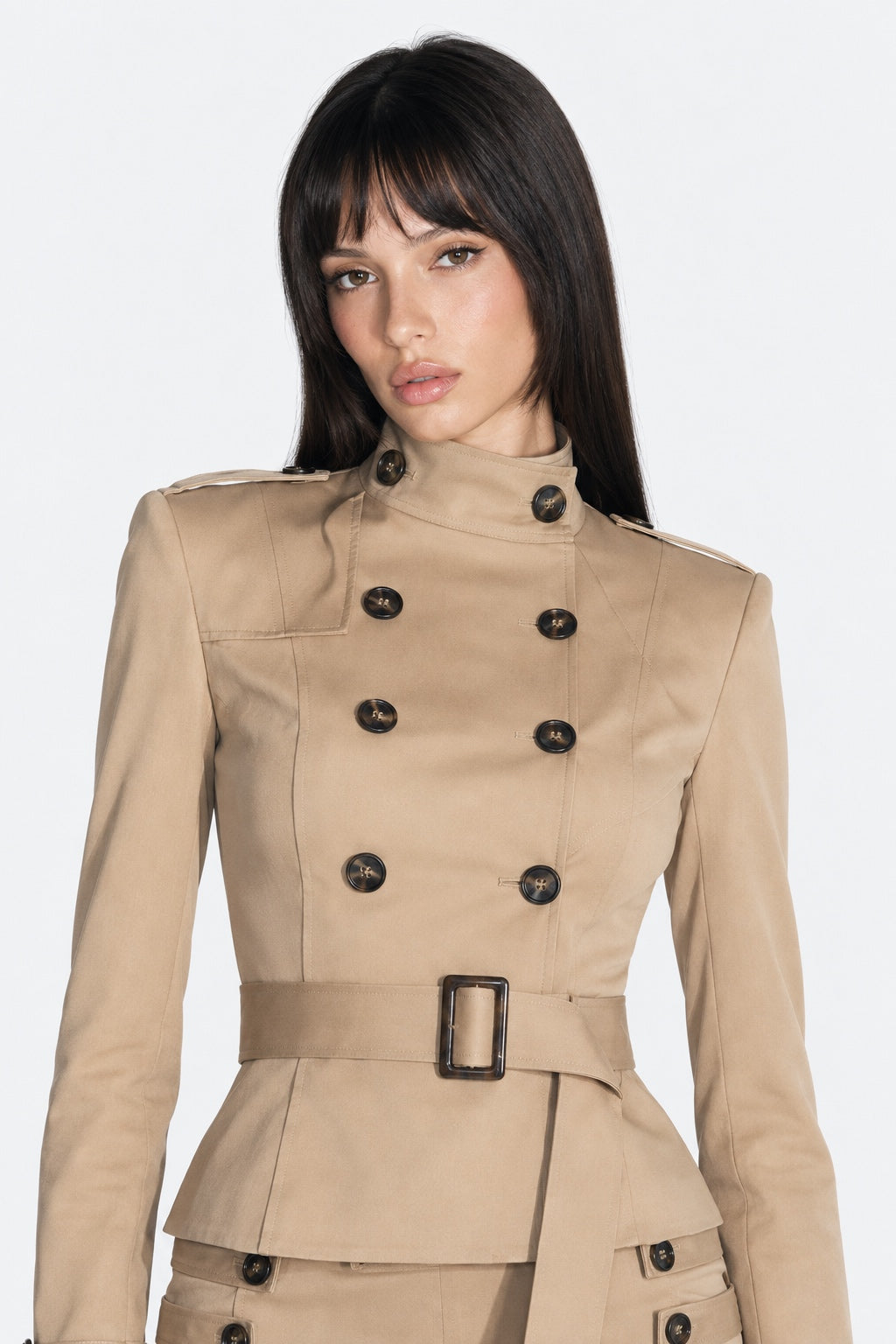 A woman modeling a beige double-breasted belted jacket with black buttons against a light gray studio background.