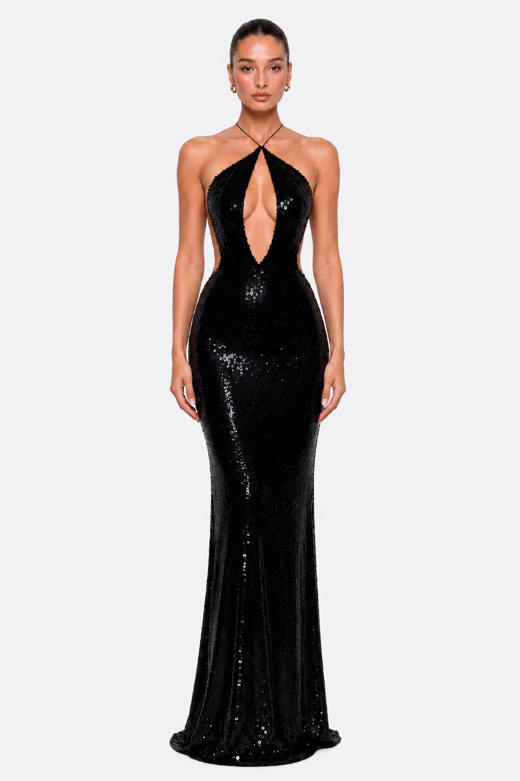A model wearing a floor-length black sequined gown with a halter neckline, plunging cutout at the bust, and a fitted silhouette that flares slightly at the hem, photographed against a light gray studio background.
