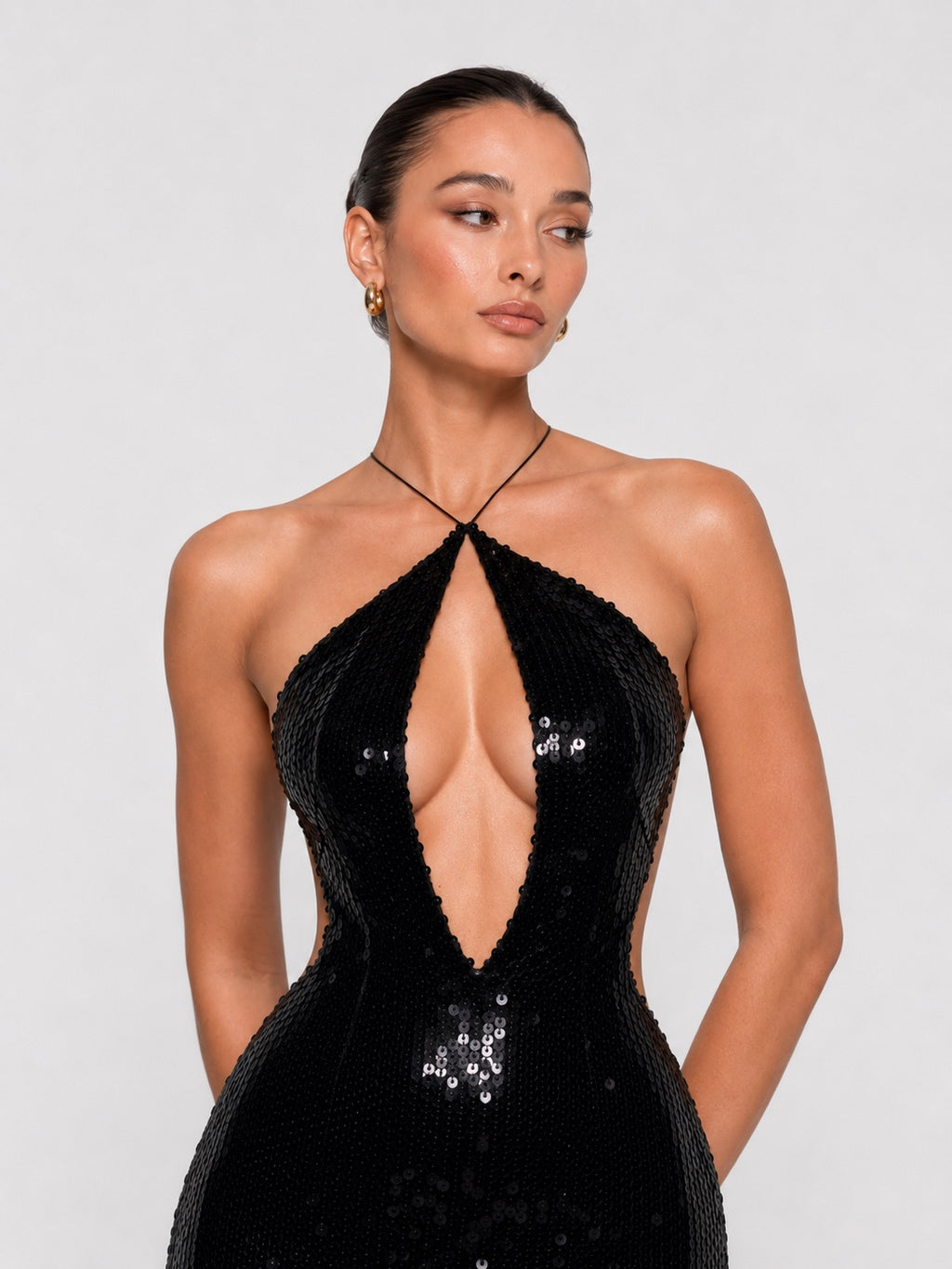 A woman modeling a black sequined halter gown with a plunging V-neckline and side cutouts, photographed against a light gray studio background.