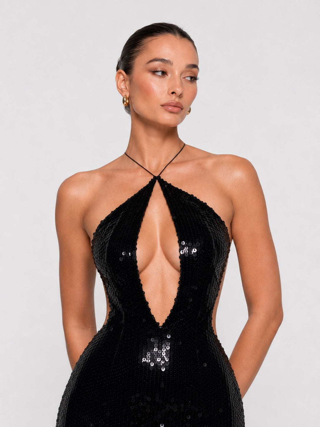 A woman modeling a black sequined halter gown with a plunging V-neckline and side cutouts, photographed against a light gray studio background.
