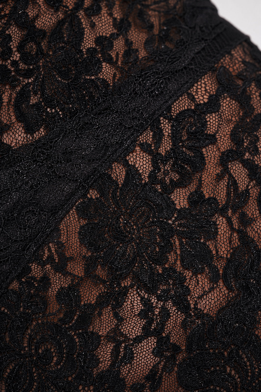Close-up of black floral lace fabric with sheer mesh showing a warm brown skin tone underneath, plus a wide black lace trim running diagonally across the frame.