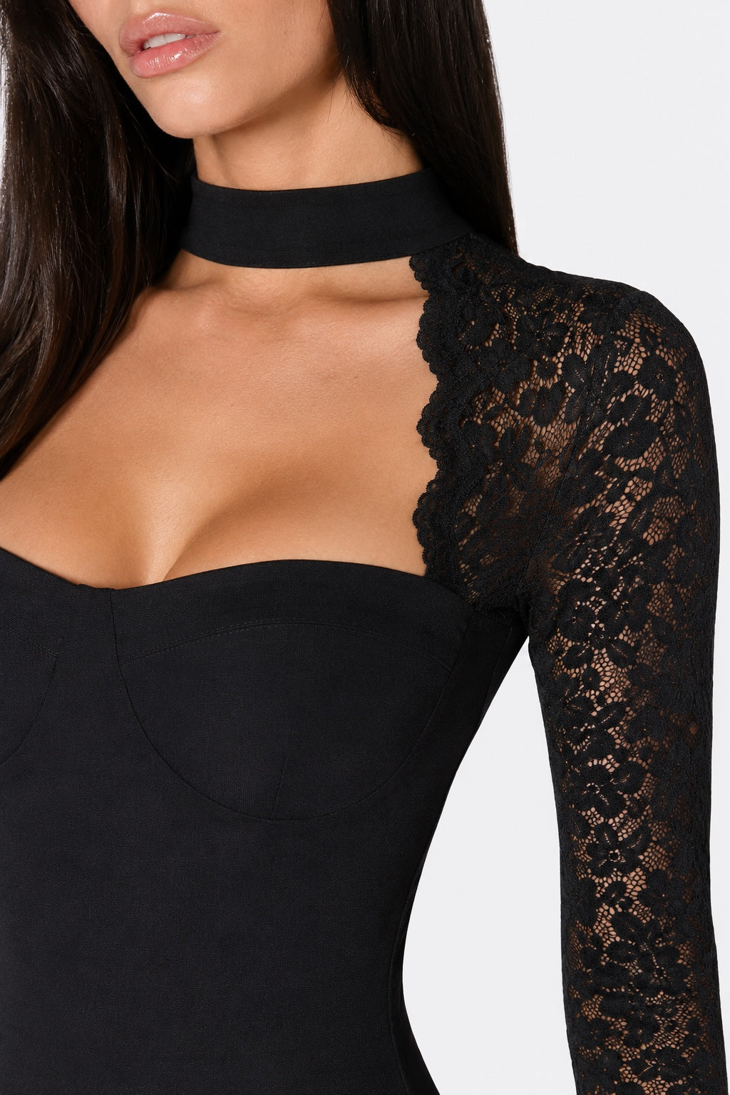Close-up of a model wearing a black long-sleeve bodysuit with a lace sleeve, a black lace choker collar, and a sweetheart neckline against a white background.