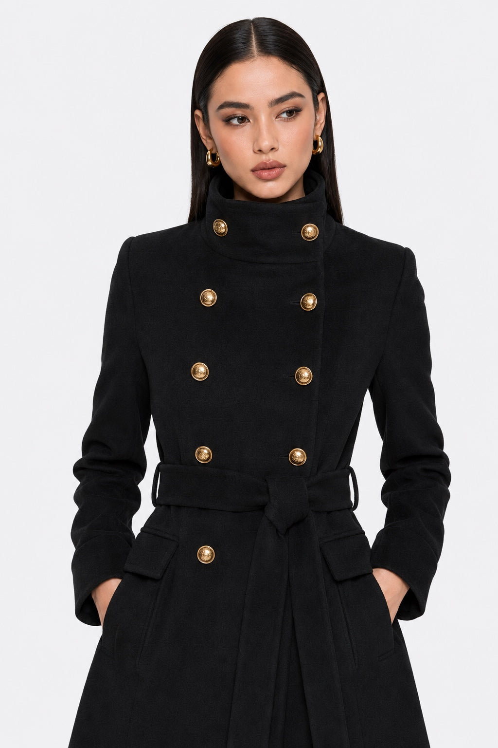 A woman modeling a black double-breasted coat with prominent gold buttons and a matching waist tie belt, set against a plain light gray studio background.
