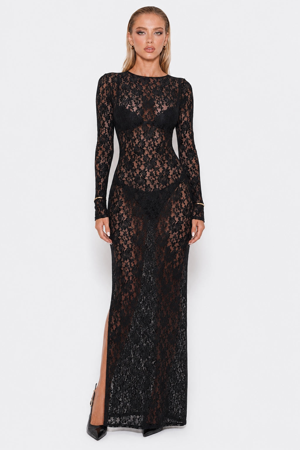 A woman modeling a long-sleeve black lace maxi dress with a thigh-high slit, photographed against a plain light gray studio background.