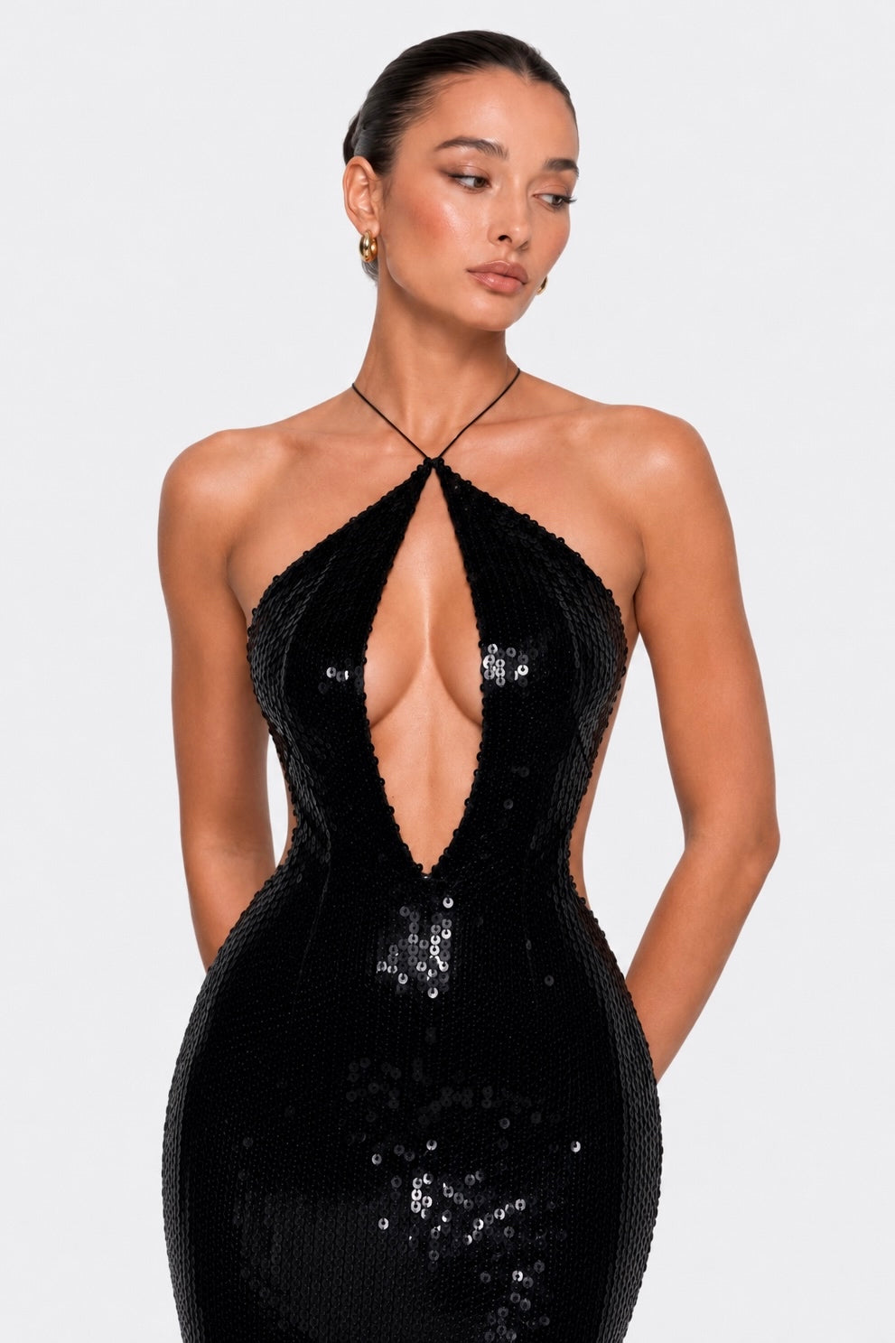A woman modeling a black sequined halter-neck dress with a plunging cutout and side cutouts, photographed against a light gray studio background.