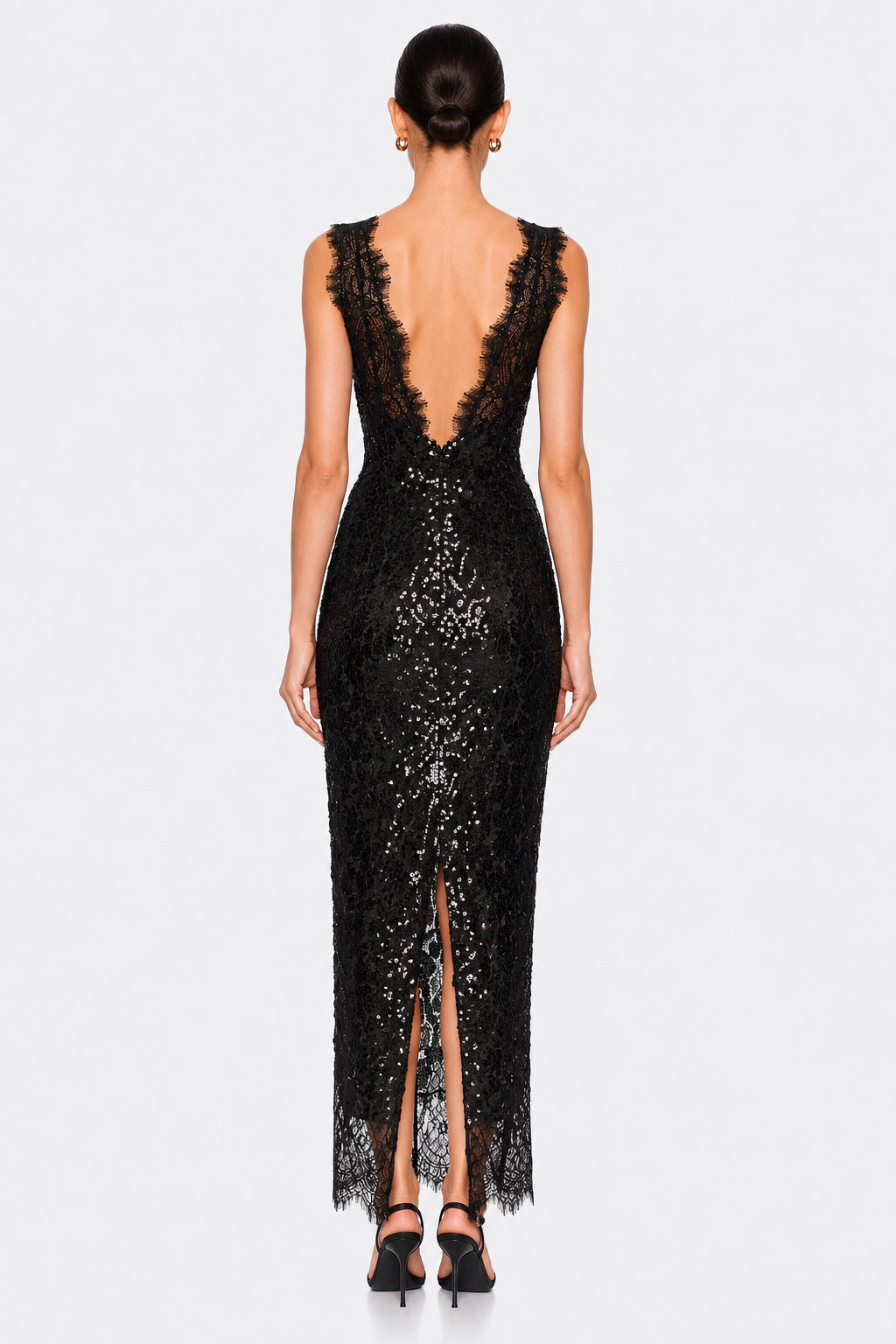 A woman shown from the back wearing a sleeveless black lace maxi dress with a deep open back, fitted silhouette, and front slit, photographed against a light gray studio background.