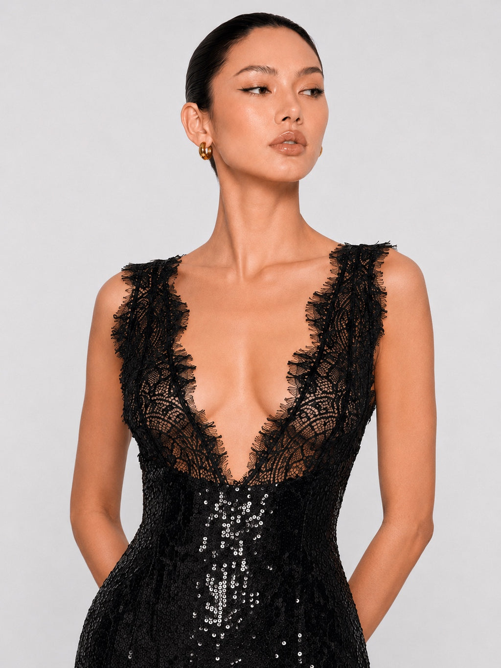 A woman modeling a sleeveless black lace and sequin gown with a deep V-neckline against a light gray studio background.