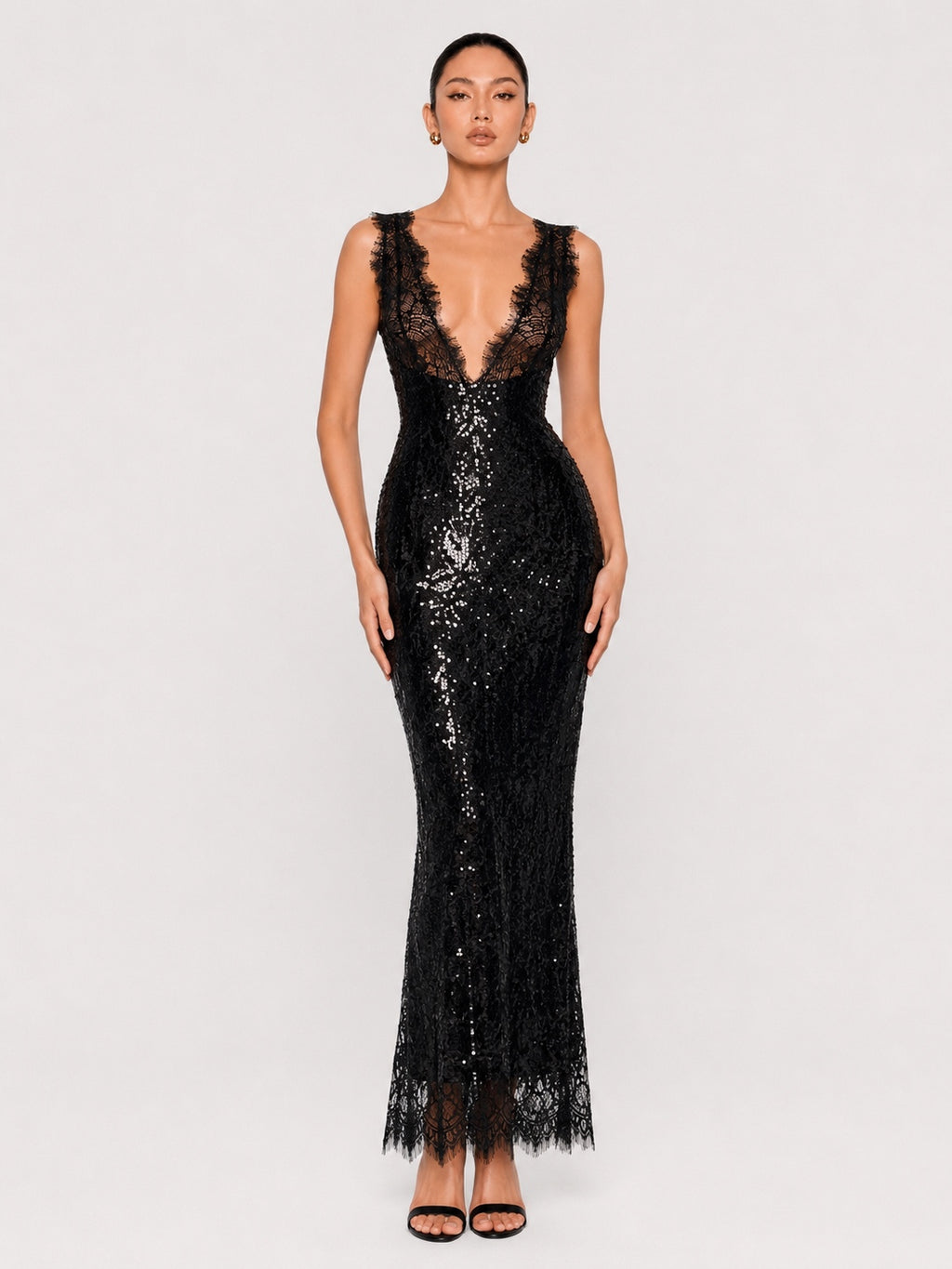 A model wearing a sleeveless black lace and sequin maxi gown with a plunging lace neckline, fitted silhouette, and lace-trimmed hem, photographed against a light gray studio background.