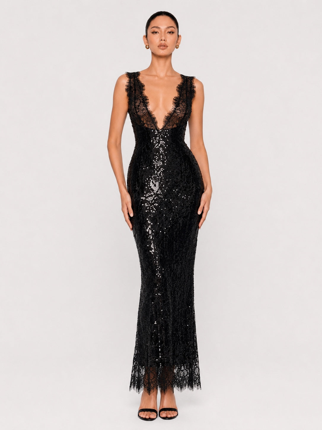 A model wearing a sleeveless black lace and sequin maxi gown with a plunging lace neckline, fitted silhouette, and lace-trimmed hem, photographed against a light gray studio background.