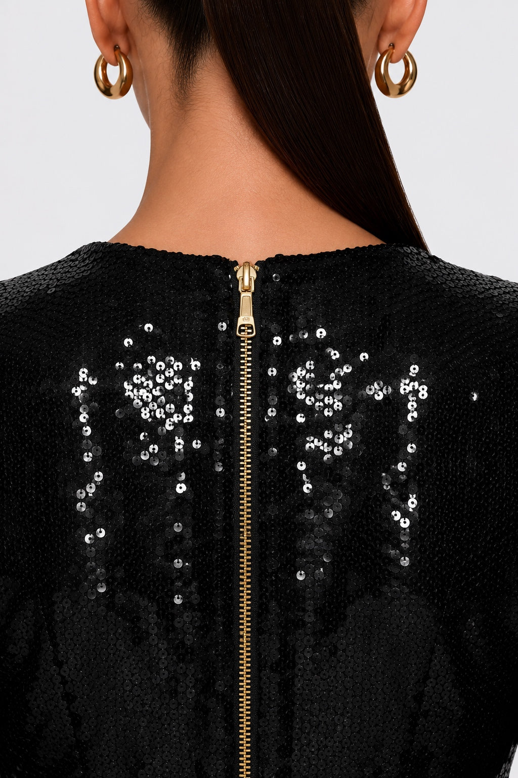 Close-up back view of a person wearing a black sequined top or dress with a gold zipper running down the center and matching gold hoop earrings.