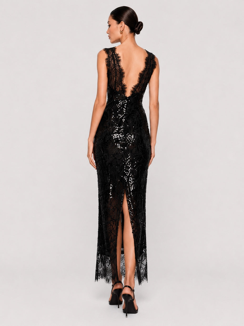 A woman modeling a black lace and sequined floor-length gown from the back, featuring a deep open back, sleeveless design, and high front slit, against a light gray studio background.