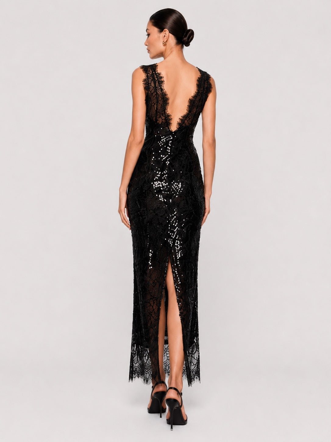 A woman modeling a black lace and sequined floor-length gown from the back, featuring a deep open back, sleeveless design, and high front slit, against a light gray studio background.