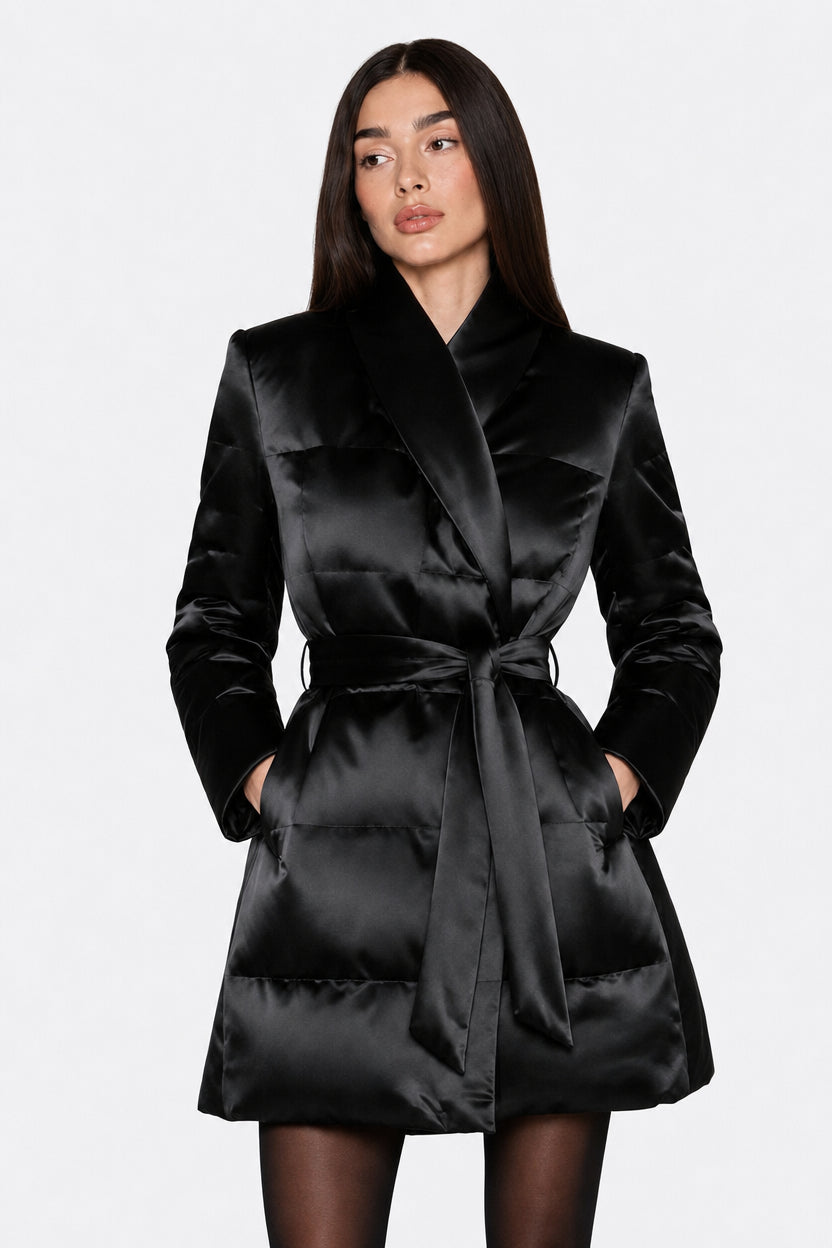A woman modeling a black satin wrap coat with a tie waist belt, photographed against a plain light gray studio background.
