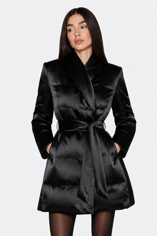 A woman modeling a black satin wrap coat with a tie waist belt, photographed against a plain light gray studio background.