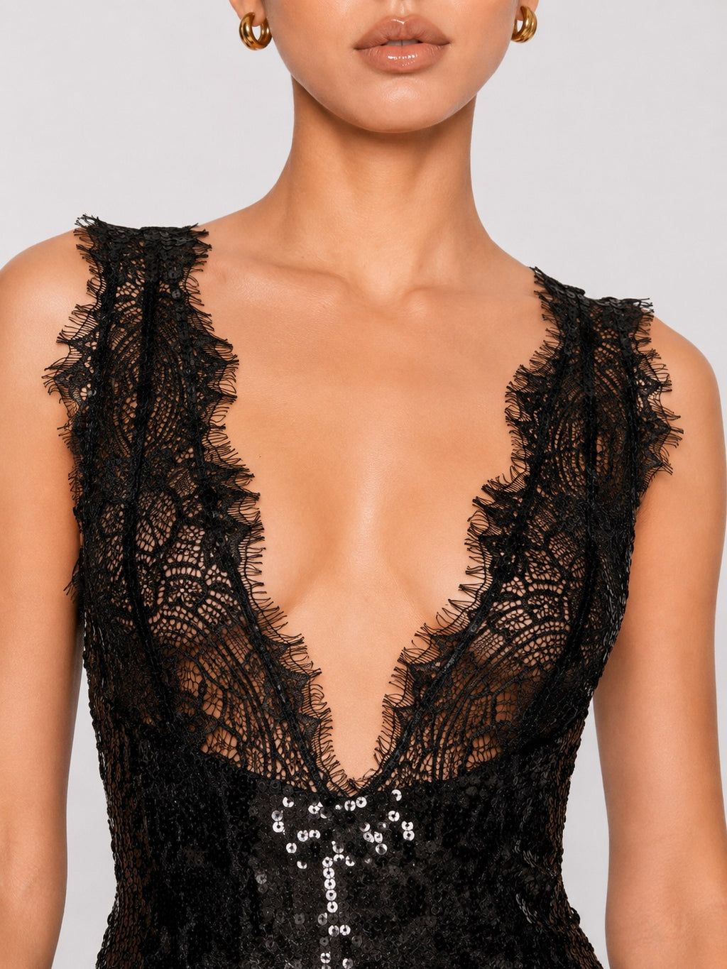 Close-up of a model wearing a black lace dress with a deep V-neckline, ruffled lace trim, and silver sequin embellishments on a light gray background.