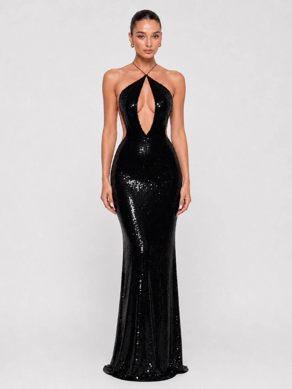 A model wearing a floor-length black sequined gown with a halter neckline, plunging keyhole cutout at the center, and a fitted silhouette that flares slightly at the bottom, photographed against a light gray studio background.