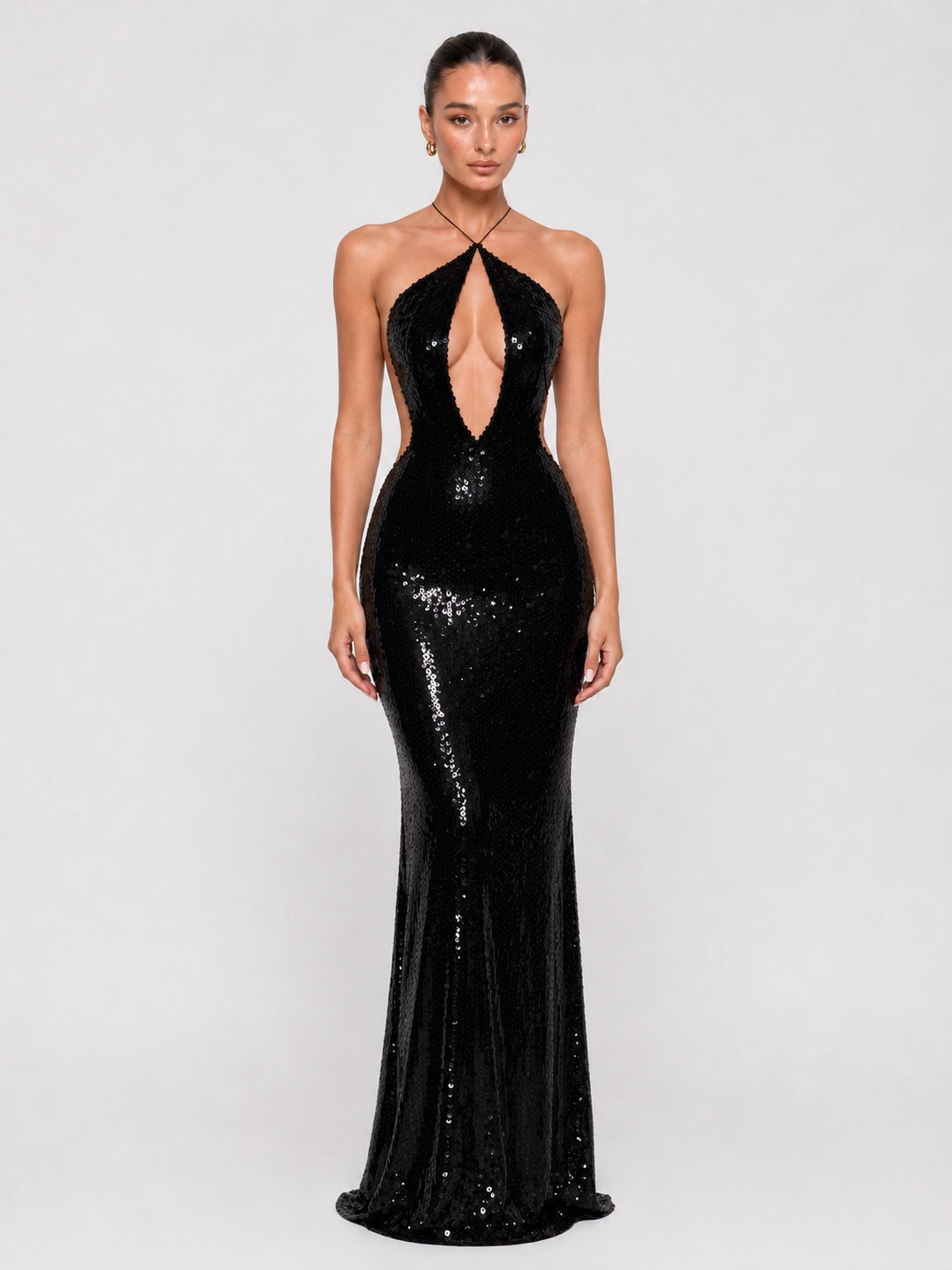 A model wearing a floor-length black sequined gown with a halter neckline, plunging keyhole cutout at the center, and a fitted silhouette that flares slightly at the bottom, photographed against a light gray studio background.