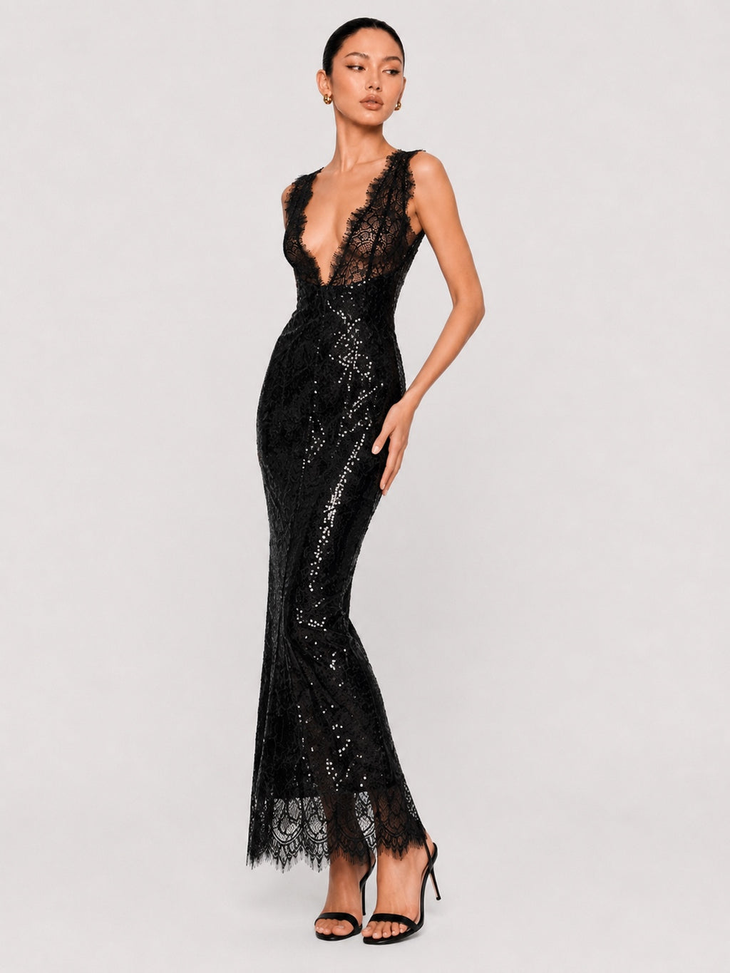 A model wearing a black lace and sequined mermaid-style evening gown with a plunging neckline, paired with black high-heeled sandals, against a light gray studio background.