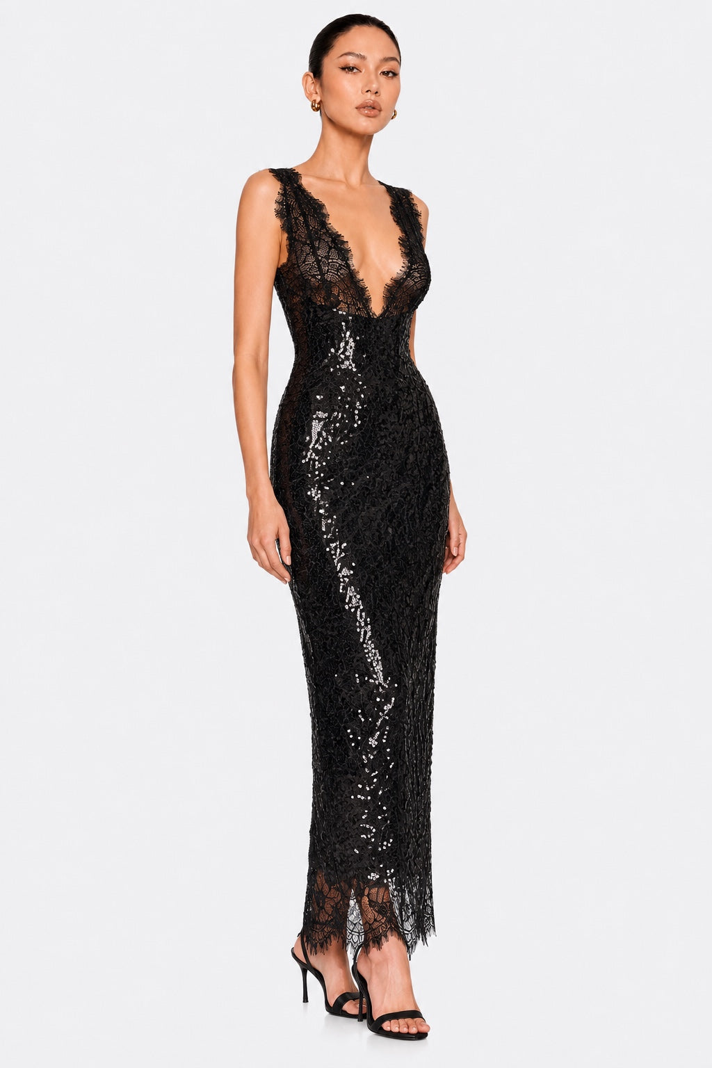 A model wearing a sleeveless black lace and sequin maxi gown with a deep V-neckline, photographed against a solid light gray background.