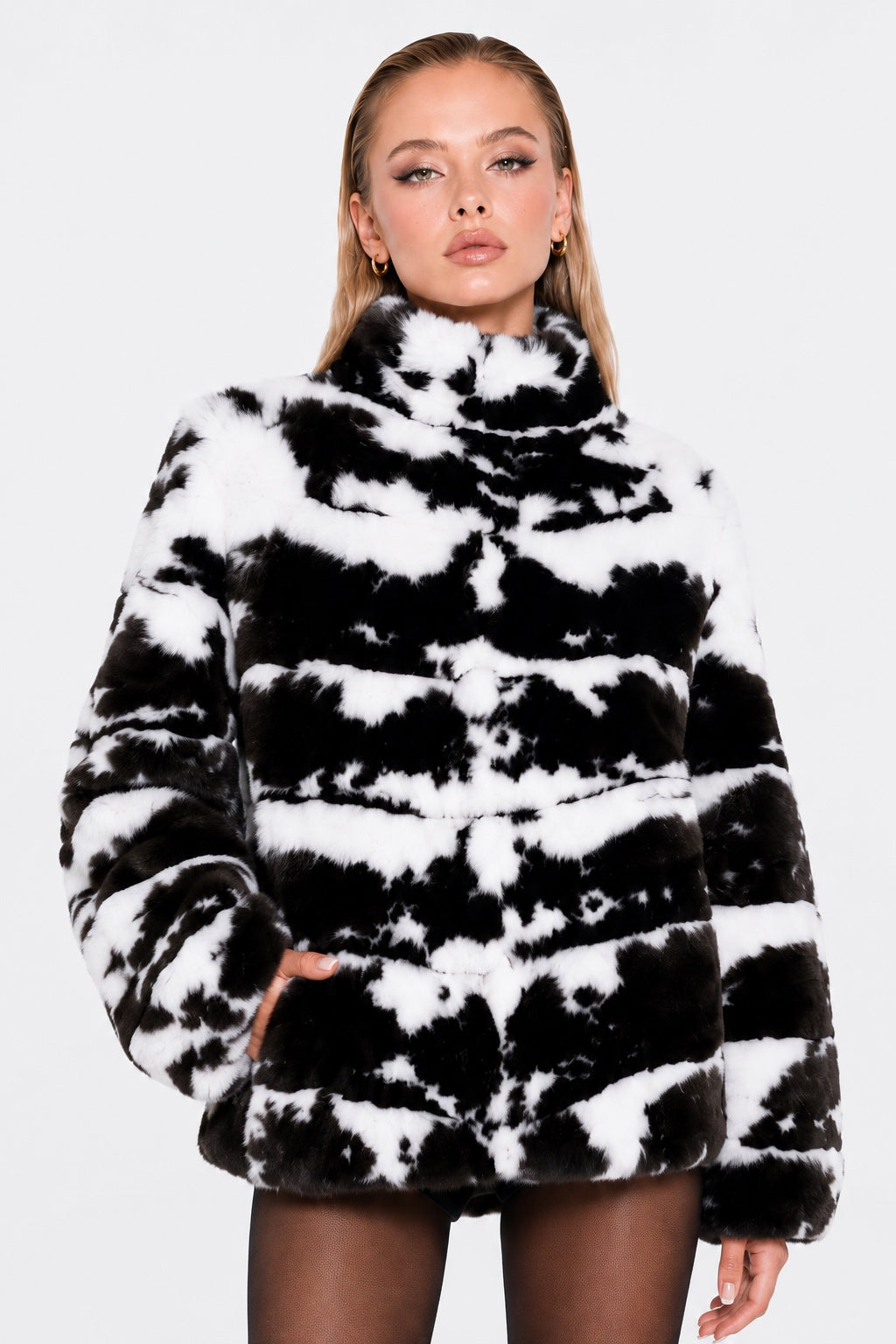 A woman modeling a black and white cow-print faux fur jacket against a light gray background.
