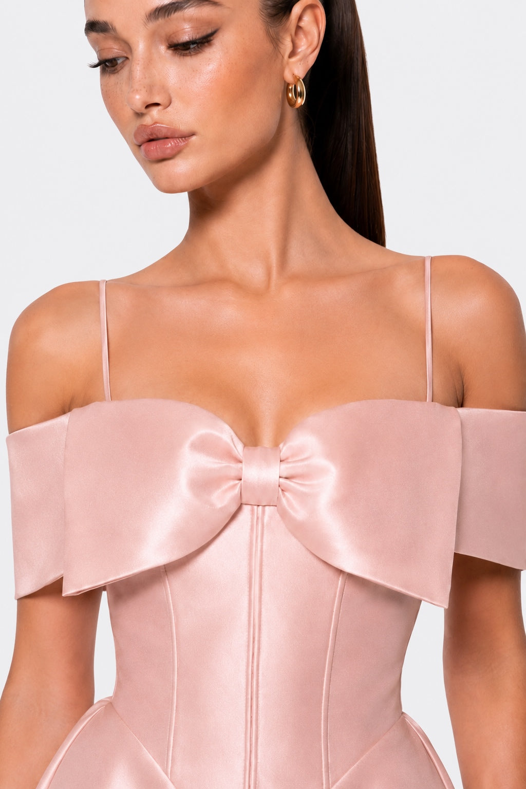 A close-up of a model wearing a pale pink satin off-the-shoulder dress with a large bow at the bust and structured corset-style bodice.