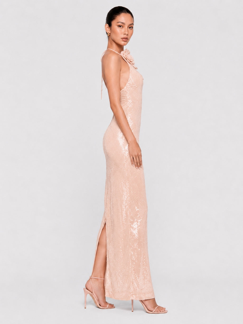 A woman modeling a floor-length pale blush pink sequined gown with a high side slit and halter-style neckline, photographed against a light gray studio background.