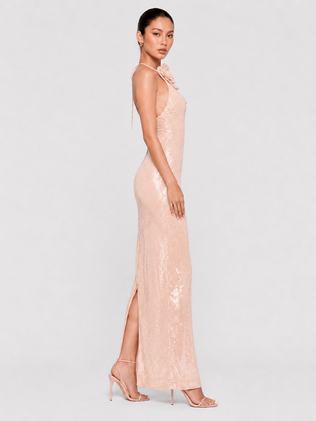 A woman modeling a floor-length pale blush pink sequined gown with a high side slit and halter-style neckline, photographed against a light gray studio background.