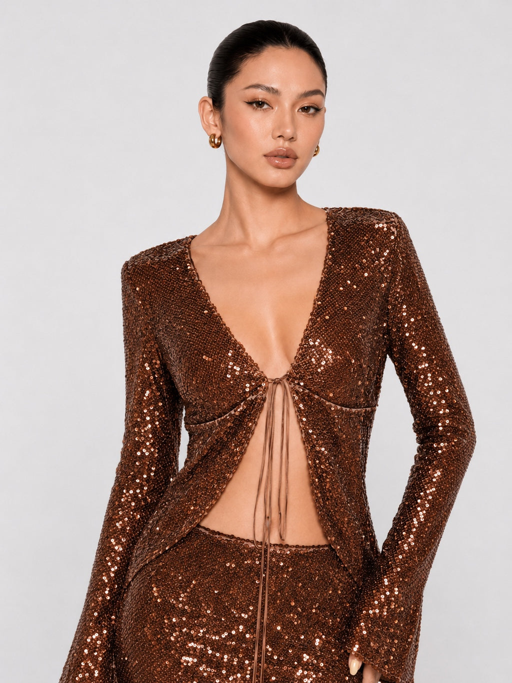 A model wearing a sparkling bronze sequined long-sleeve top with a deep V-neckline and front tie detail, paired with matching sequined bottoms, against a light gray studio background.