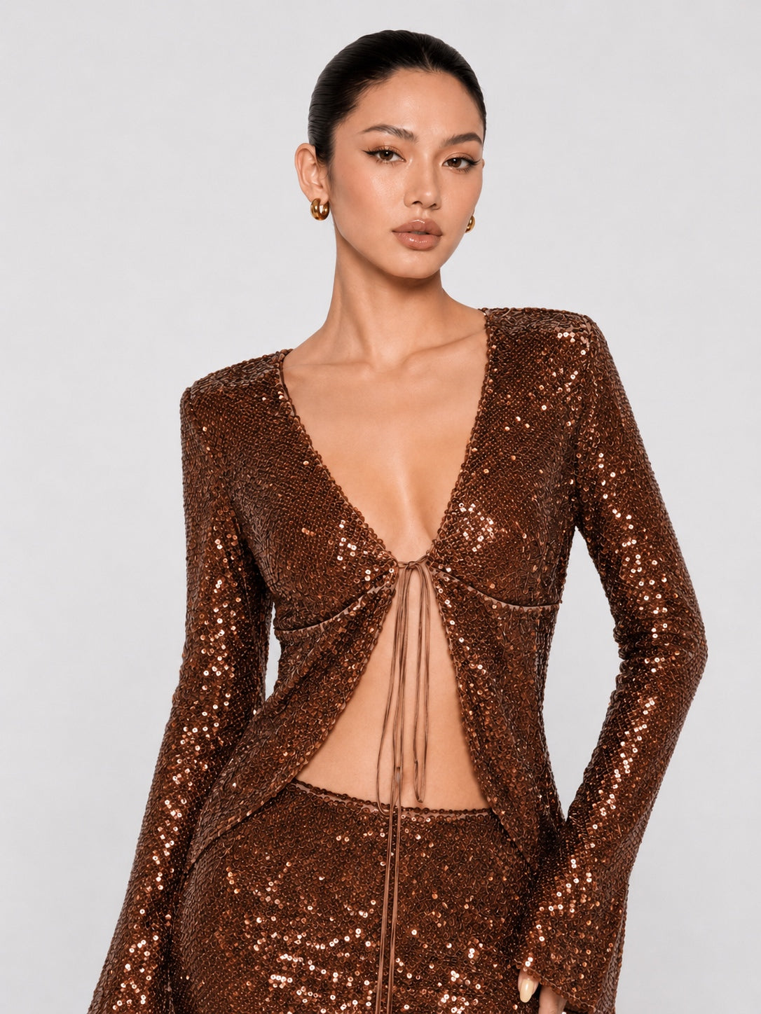 A model wearing a sparkling bronze sequined long-sleeve top with a deep V-neckline and front tie detail, paired with matching sequined bottoms, against a light gray studio background.