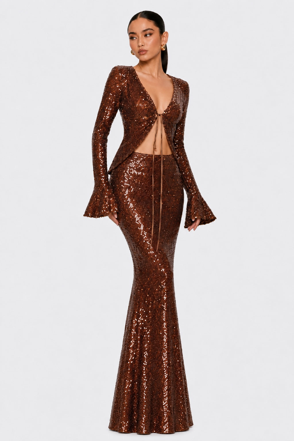 A model wearing a long-sleeved, deep V-neck, floor-length sequin gown in a metallic bronze-brown color, set against a plain light gray background.