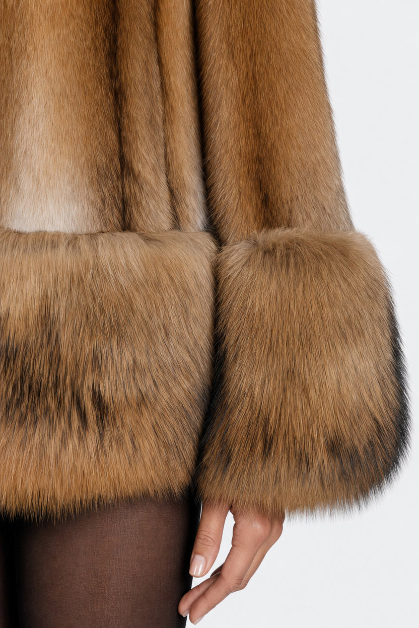 Close-up of a brown fur jacket with wide cuffs, worn by a model whose hand and legs are visible against a plain light background.