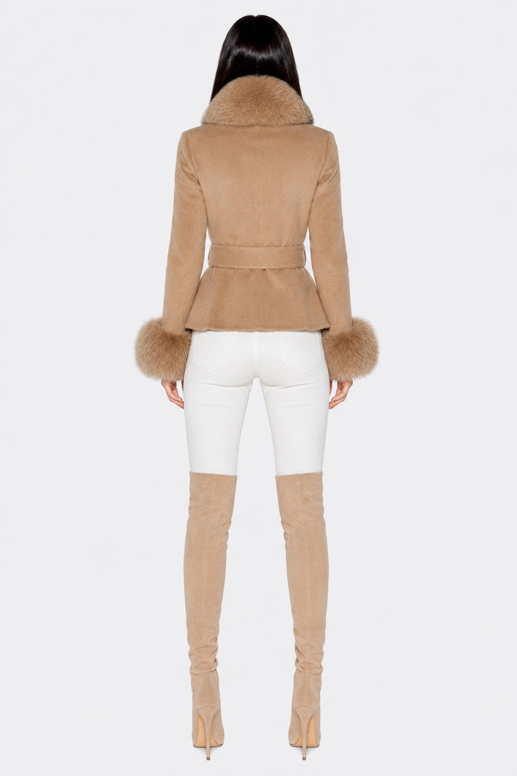 A model shown from the back wearing a tan shearling jacket with a fur collar and cuffs, paired with white skinny pants and matching tan over-the-knee boots, against a plain white background.