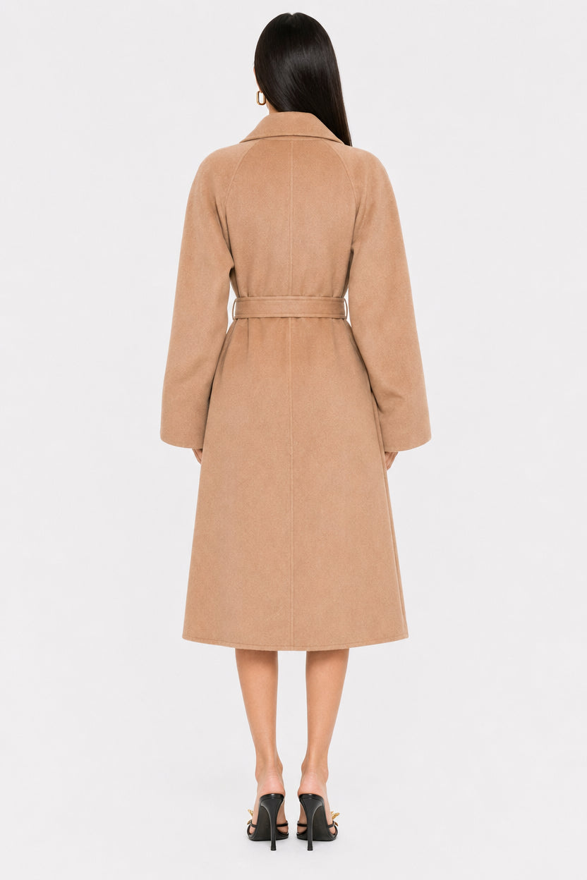 A woman viewed from the back wearing a camel-colored belted coat and black heeled sandals against a plain light gray studio background.