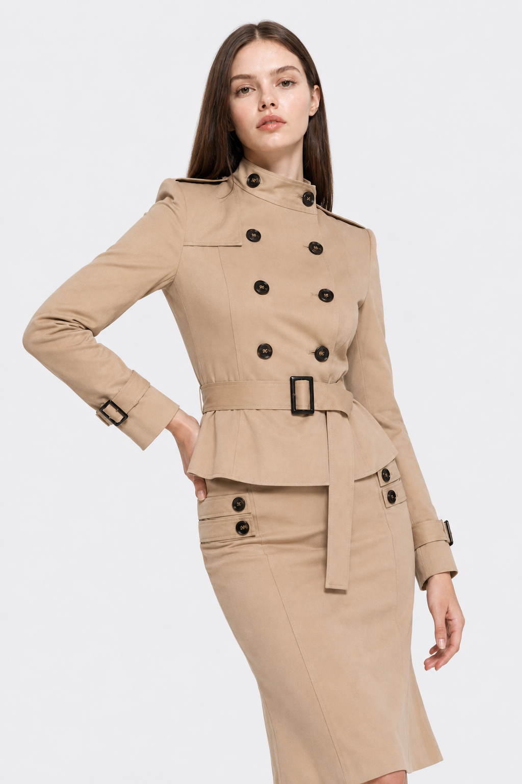 A woman modeling a beige double-breasted belted jacket and matching pencil skirt set with black buttons, against a light gray studio background.