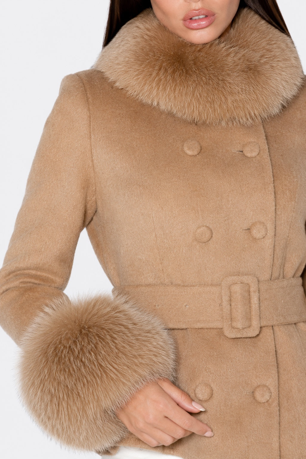 A close-up of a woman wearing a camel-colored double-breasted coat with a large fluffy faux-fur collar and matching fur-trimmed cuffs, against a plain light background.