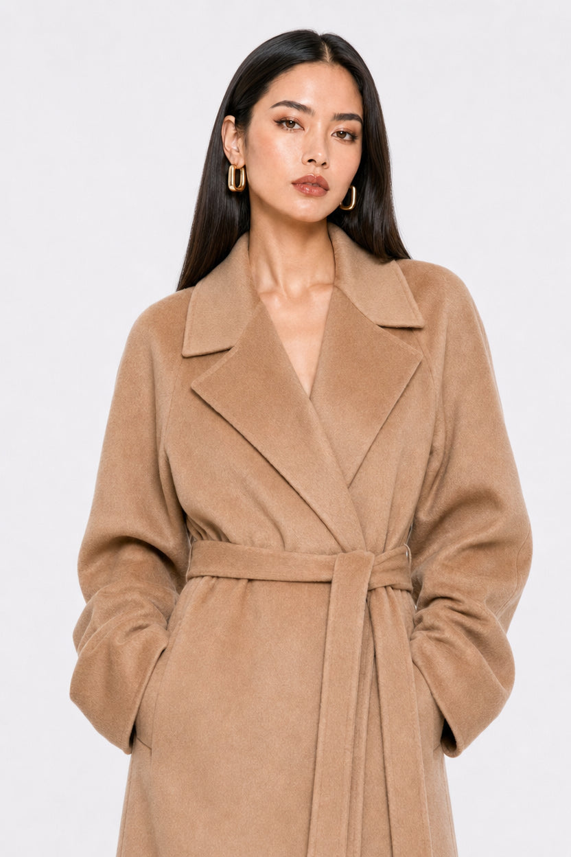 A woman modeling a camel-colored wool wrap coat with a matching waist belt, set against a plain light gray studio background.