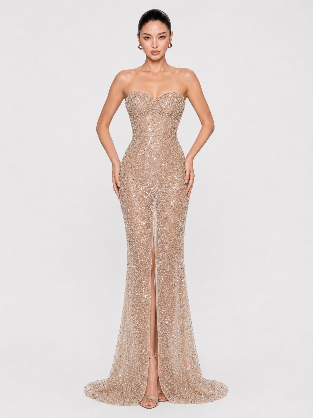 A woman modeling a strapless champagne sequined gown with a sweetheart neckline, fitted silhouette, thigh-high slit, and floor-length train against a light gray studio background.