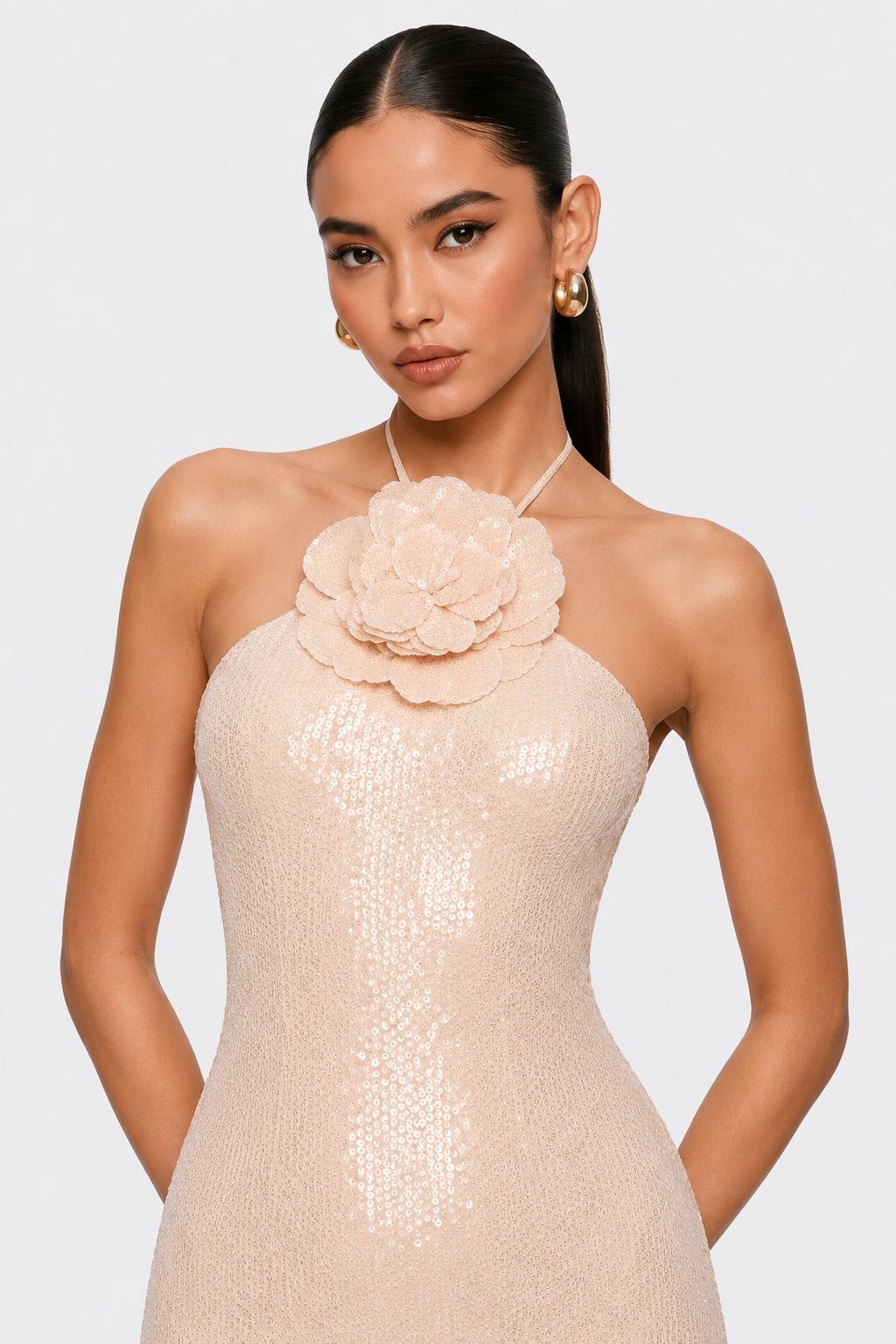 A woman modeling a champagne-colored halter dress with a large 3D flower applique at the neckline and shimmering sequins, set against a plain light gray studio background.