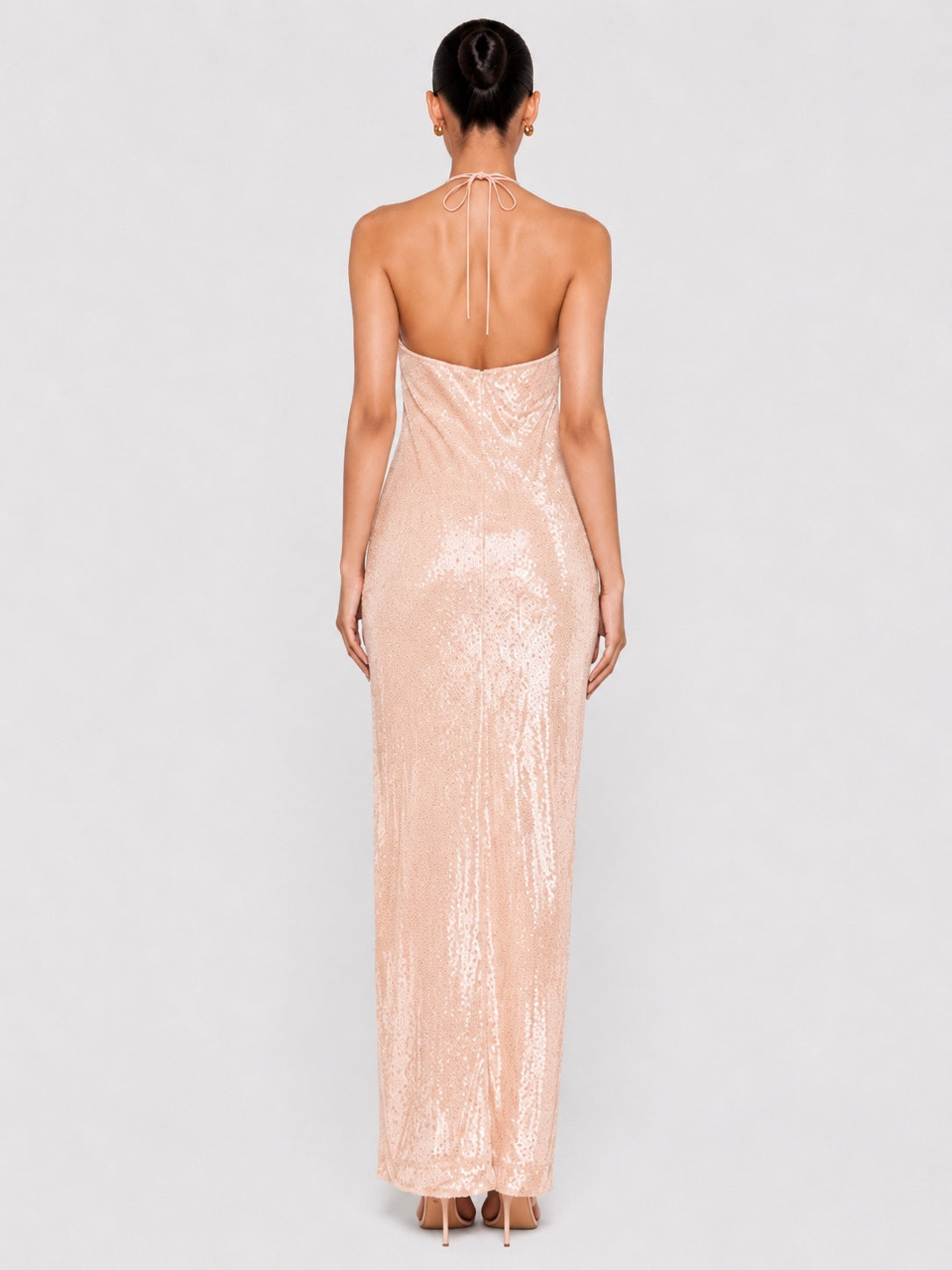 A woman viewed from the back wearing a sleeveless, backless champagne pink sequined maxi dress against a light gray studio background.