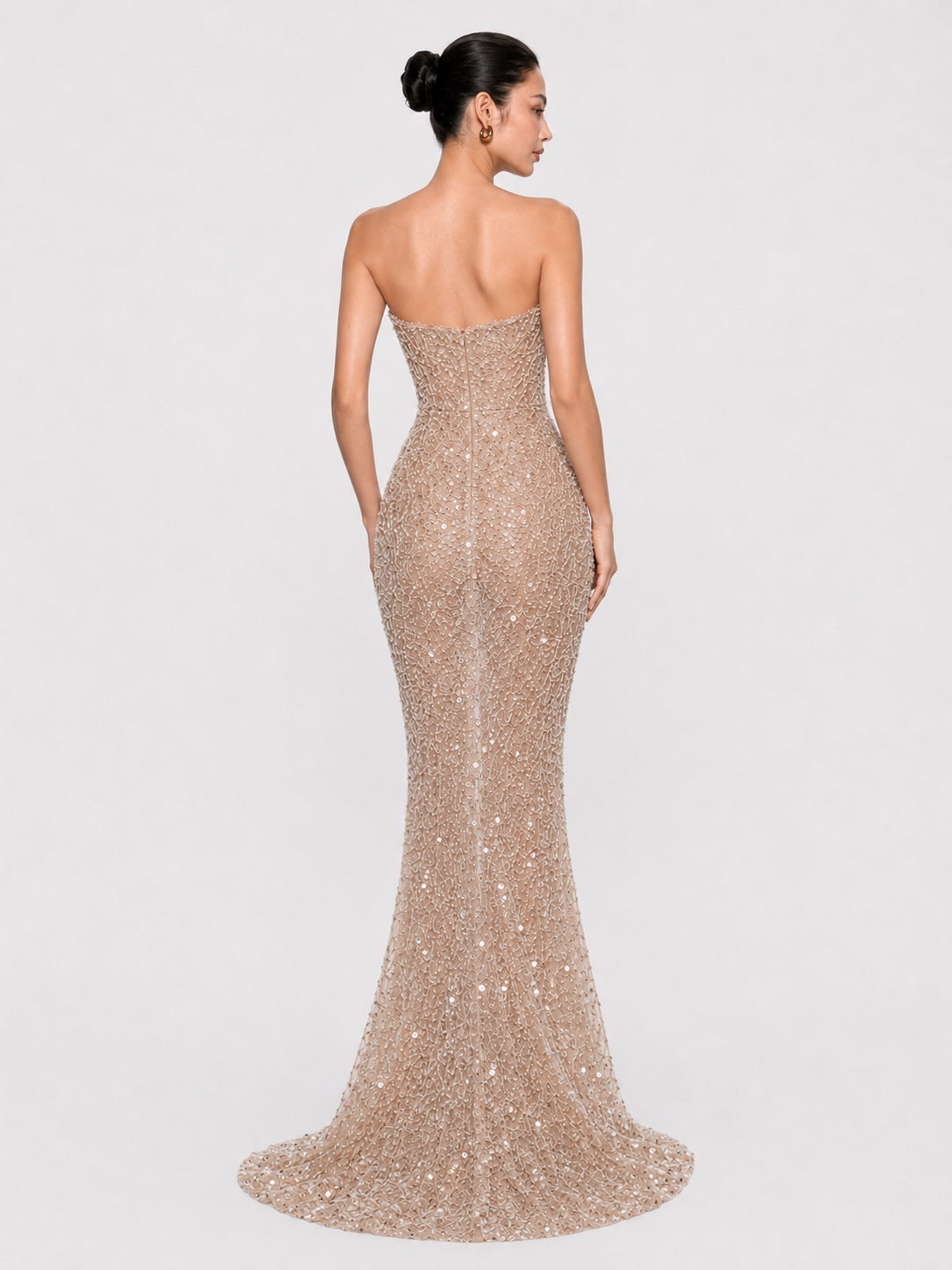A woman modeling a strapless, floor-length mermaid gown covered in shimmering sequins, shown from the back against a light gray studio background.