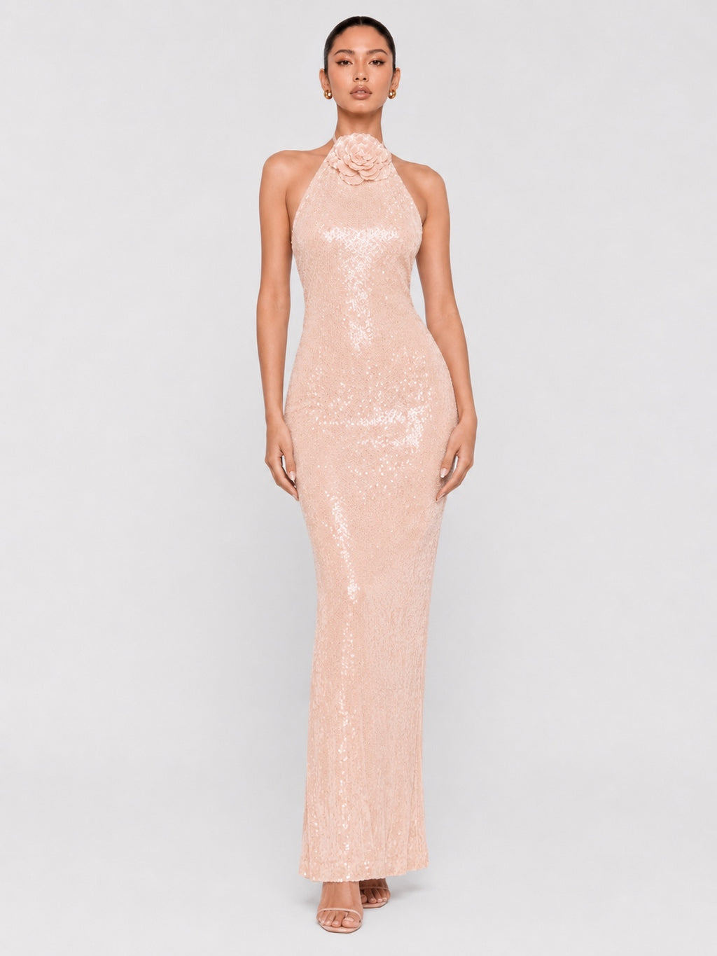 A model wearing a pale pink sequined halter-neck maxi dress with a floral embellishment at the neckline, set against a plain light gray studio background.