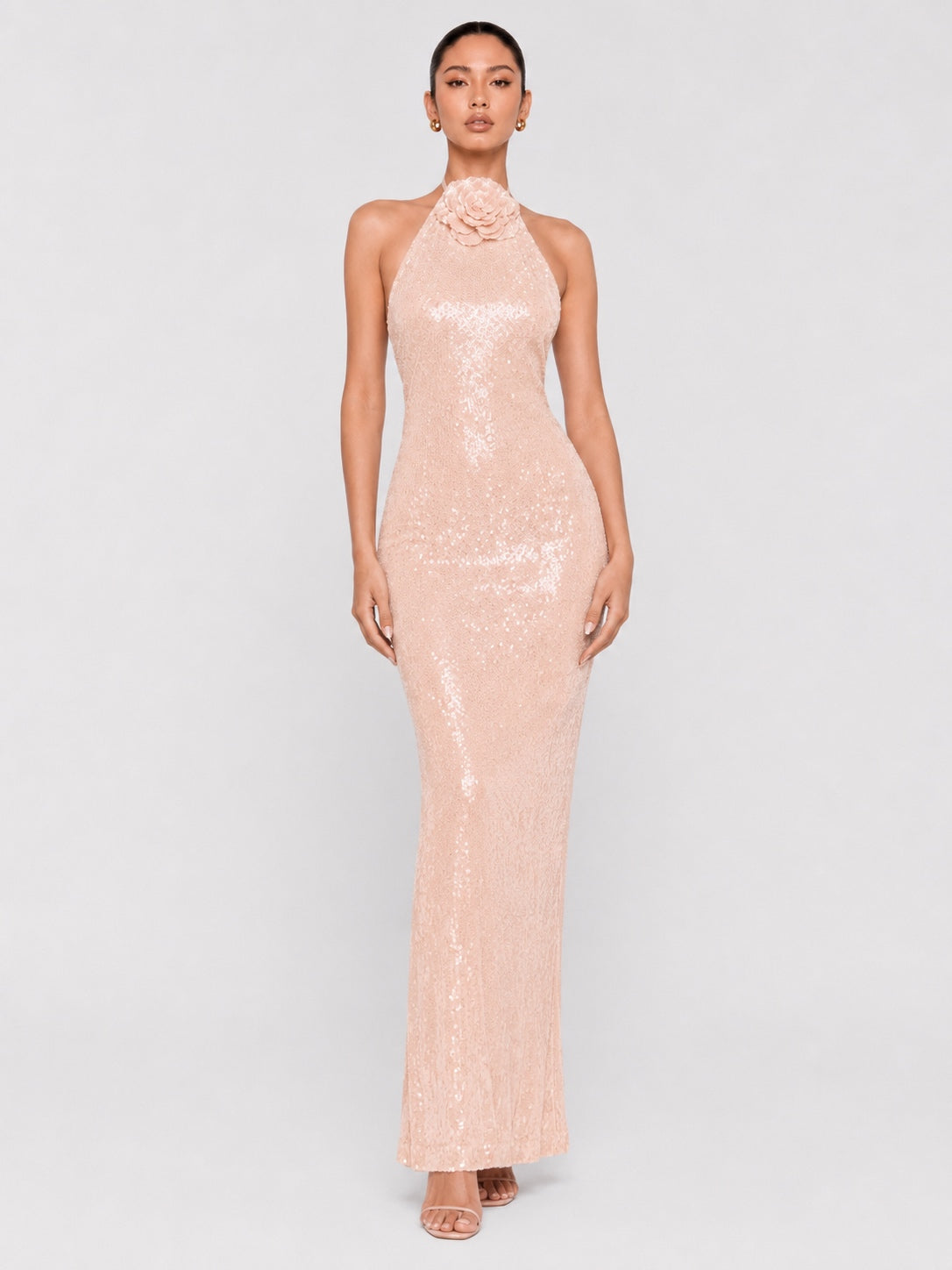 A model wearing a pale pink sequined halter-neck maxi dress with a floral embellishment at the neckline, set against a plain light gray studio background.