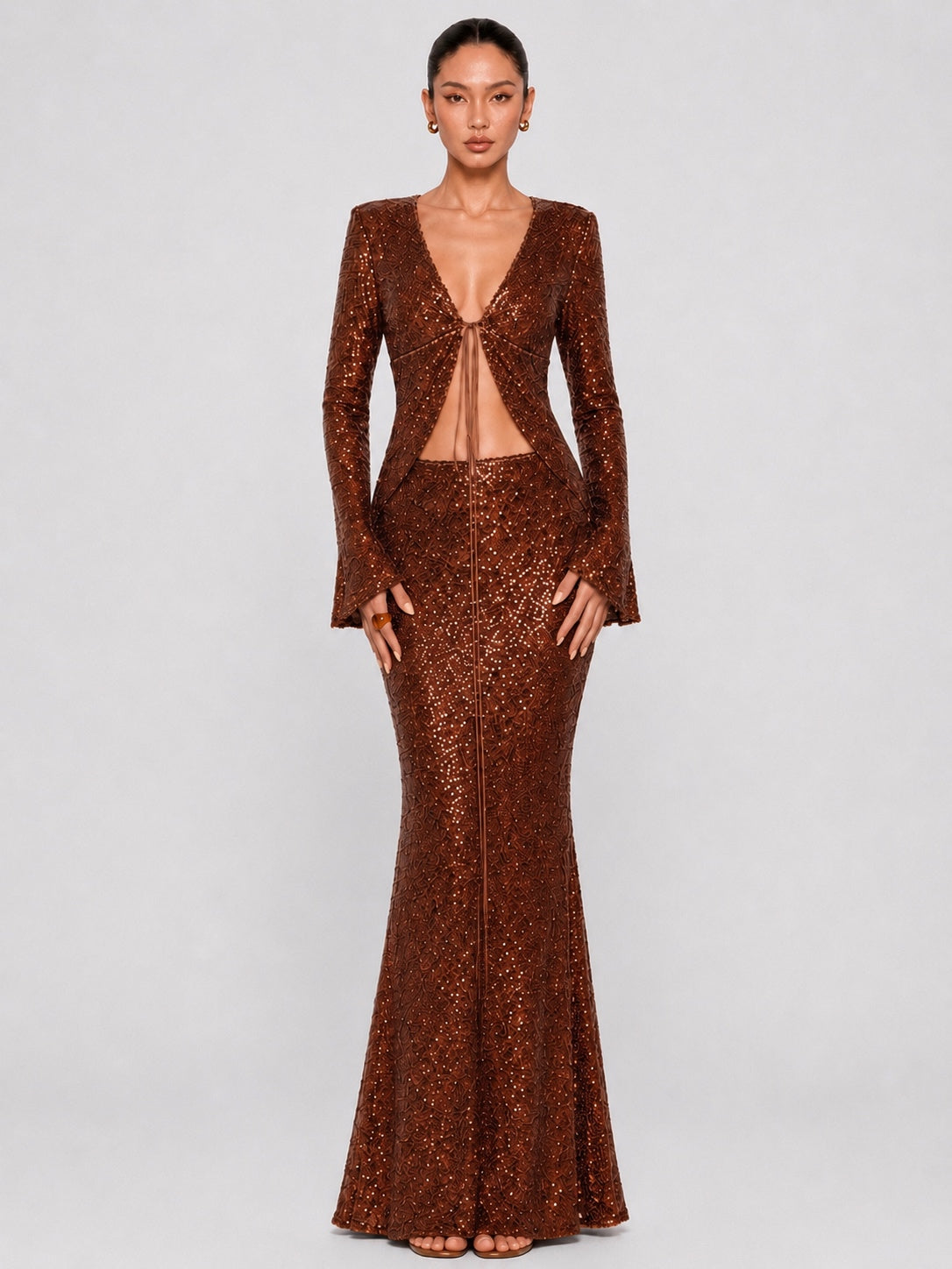 A model wearing a floor-length, sparkly bronze-brown sequined gown with long sleeves, a deep V-neckline, and a front cutout, photographed against a light gray studio background.