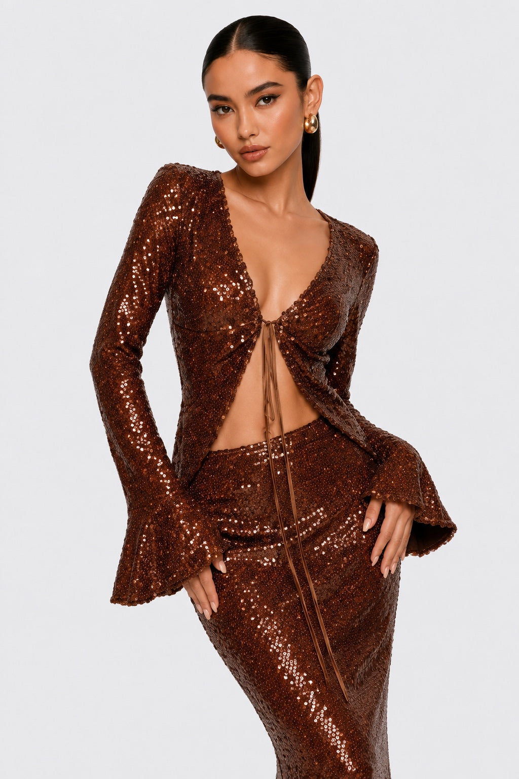 A woman modeling a sparkling bronze sequined two-piece outfit with a long-sleeve wrap-style top and matching fitted skirt against a light gray studio background.