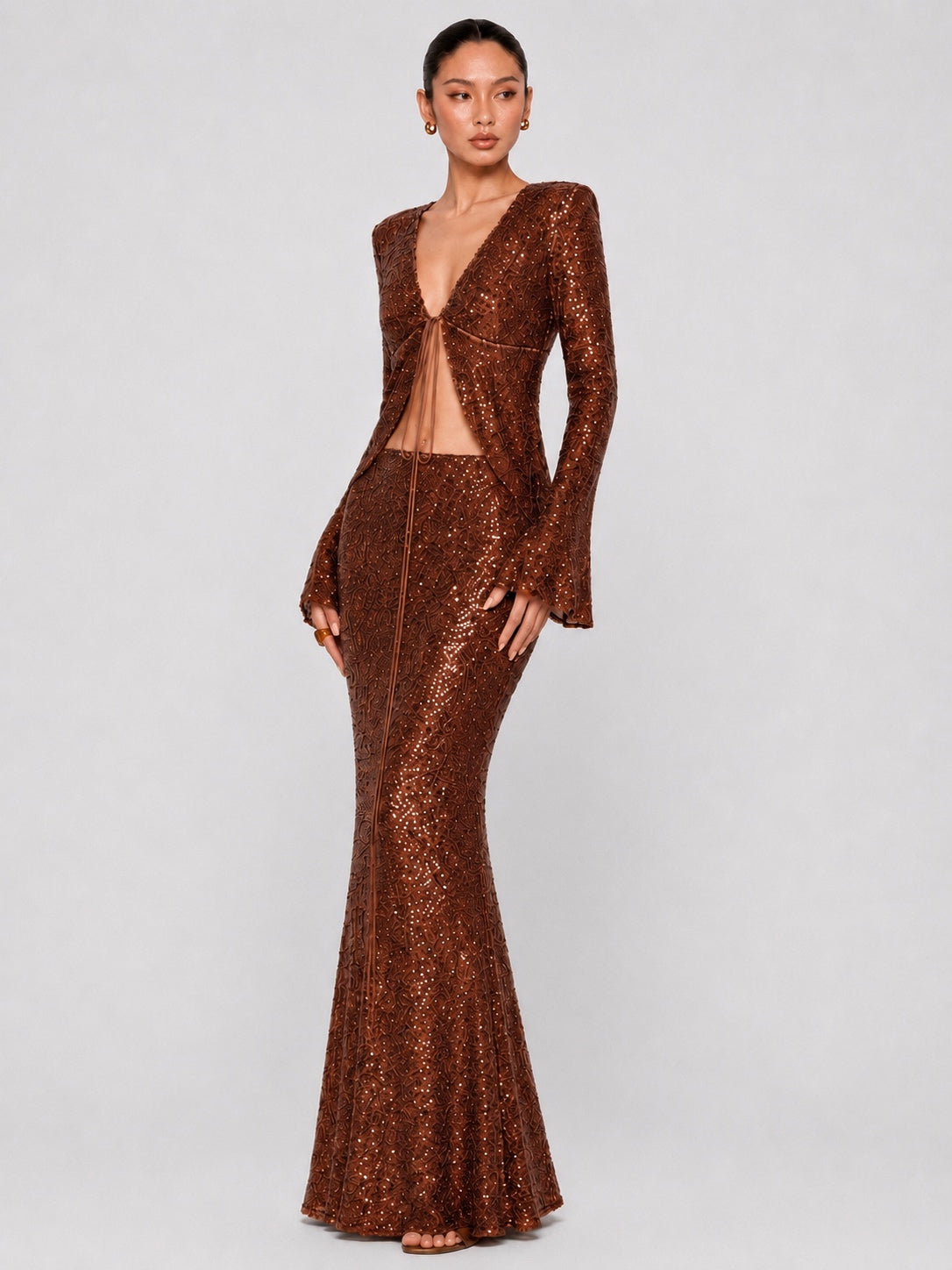 A model wearing a sparkling bronze sequined two-piece set consisting of a long-sleeve cutout top and a matching fitted mermaid skirt, photographed against a neutral light gray studio background.