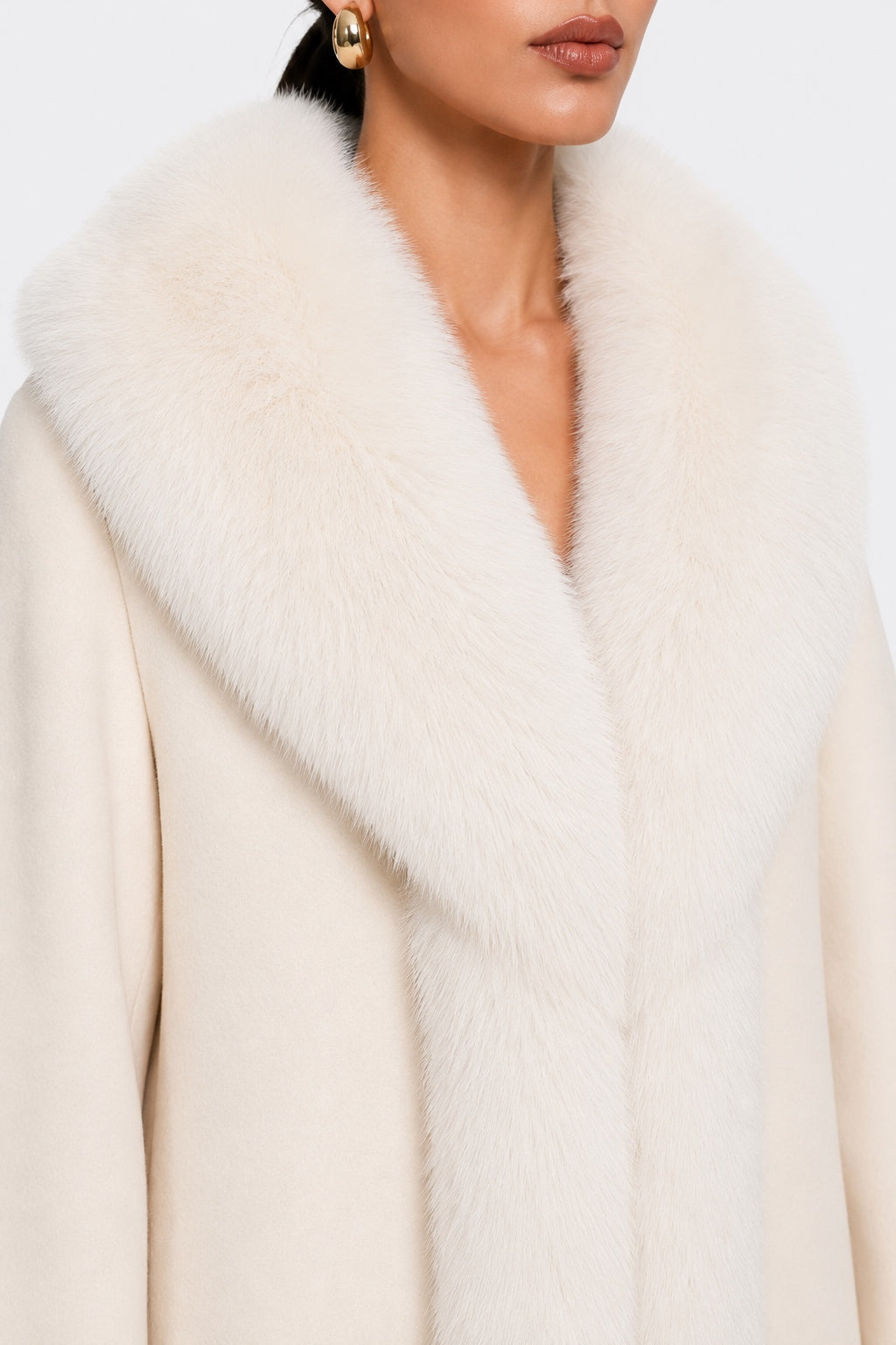 A close-up of a model wearing a luxurious cream-colored coat with a thick, fluffy natural-fur collar and a smooth cream body, set against a plain light background.