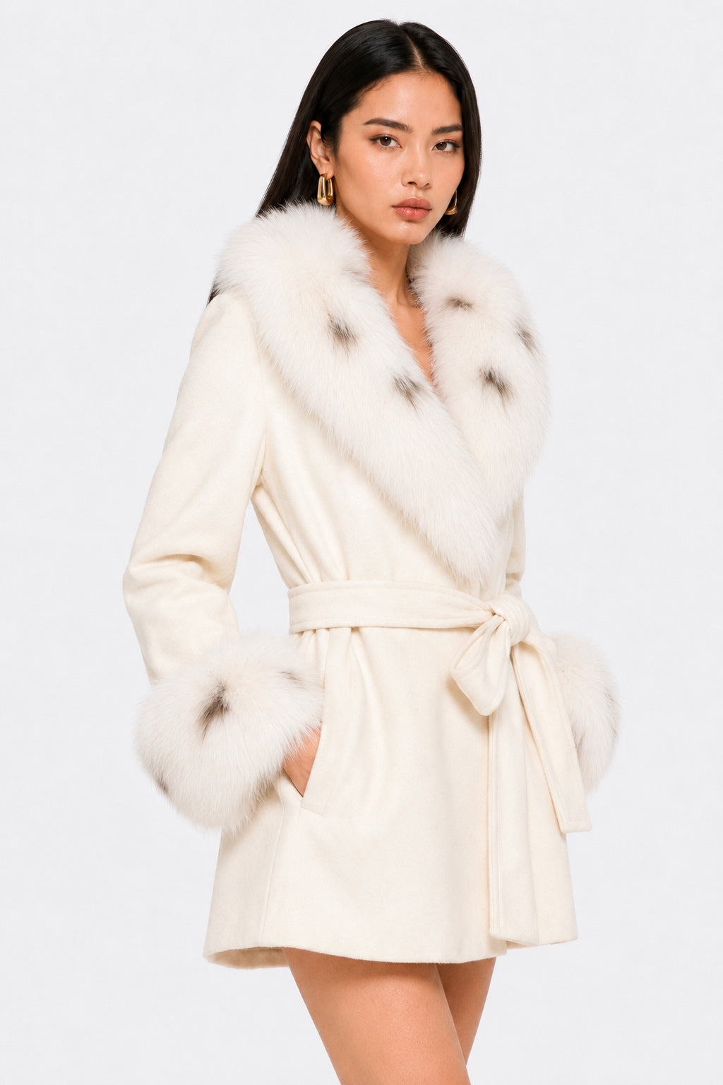 Model wearing an ivory 100% wool belted coat with a large plush fox fur collar and matching fox fur cuffs, shown in a three-quarter front pose with both hands in the pockets