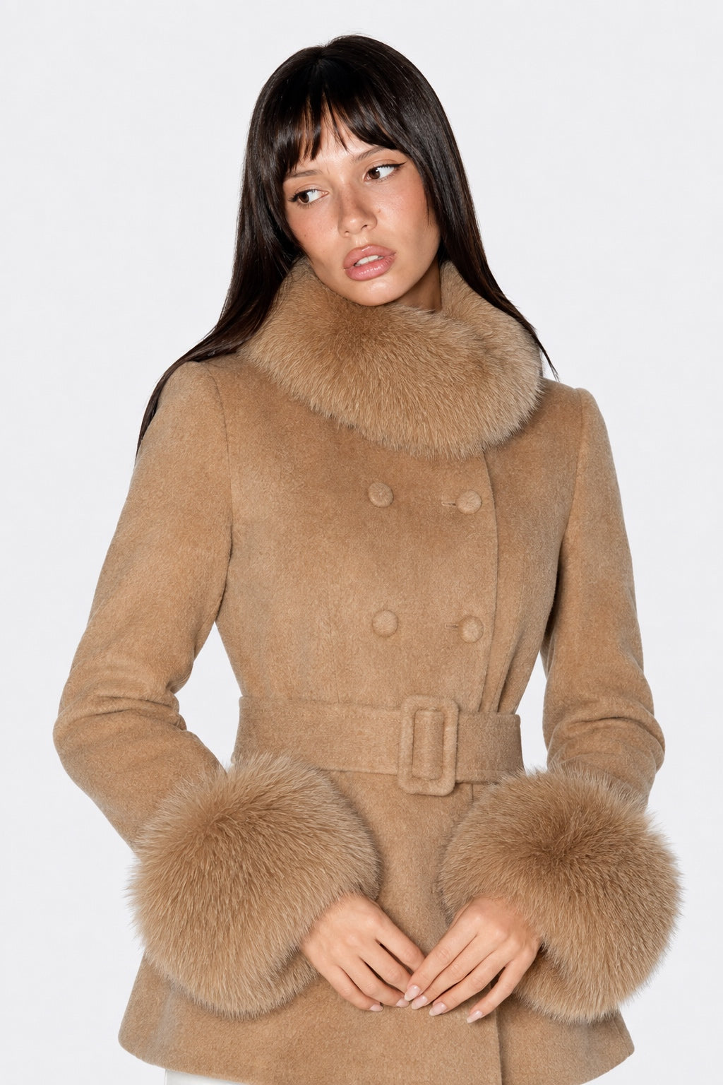 A woman wearing a camel-colored double-breasted coat with a large fur collar, matching belt, and fluffy fur cuffs, posed against a plain white studio background.