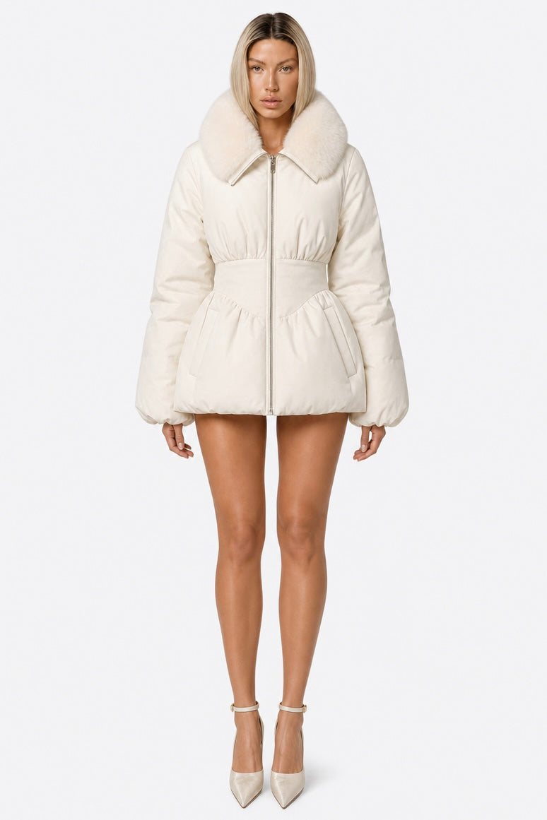 A model wearing a cream-colored puffer jacket with a large faux-fur collar, a zippered front, and a peplum waist, paired with beige high heels against a plain white background.