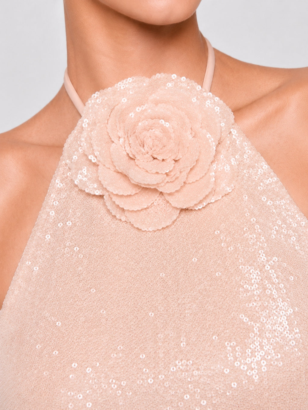 Close-up of a model wearing a pale blush pink halter top with a large layered fabric flower applique on the chest and scattered sequins across the fabric.